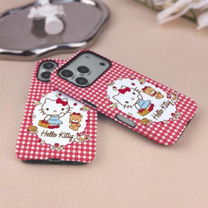 May include: Two Hello Kitty phone cases. The cases have a red and white gingham pattern. Each case features a Hello Kitty graphic with a teddy bear and the words "Hello Kitty".