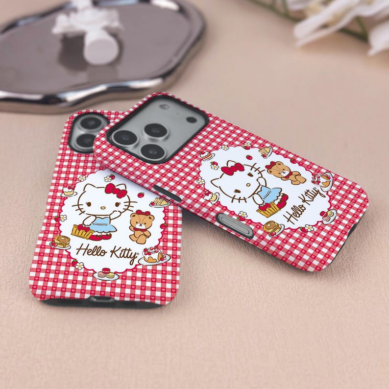 May include: Two Hello Kitty phone cases. The cases have a red and white gingham pattern. Each case features a Hello Kitty graphic with a teddy bear and the words "Hello Kitty".