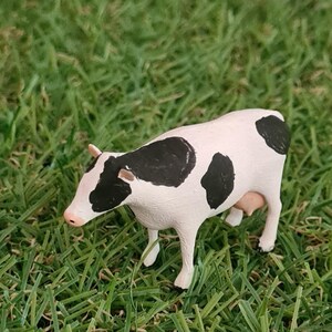 May include: A small, white cow figurine with black spots stands on green grass. The miniature cow has a pink nose and udders. The background includes a miniature house and red fence.