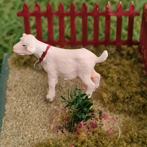 May include: A miniature goat figurine with a red collar stands within a small enclosure. The enclosure features a red picket fence and a base covered in green and tan textured materials, simulating grass and dirt. The background is a blurred green.