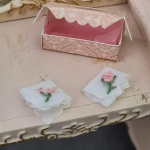 May include: A miniature vanity scene featuring a small pink and white box with a scalloped edge, and two white handkerchiefs with embroidered pink roses. The scene is set on a light pink surface with a decorative mirror and table.