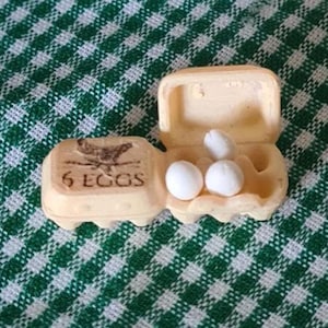 May include: A miniature egg carton with the text "6 EGGS" and a hen illustration. The carton is open, revealing three white eggs. The carton is beige and sits on a green and white checkered cloth.