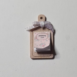 Puede incluir: Una tabla de cortar de madera en miniatura con un pequeño bloc de notas que dice "Shopping List". El bloc de notas está atado con una cinta morada.