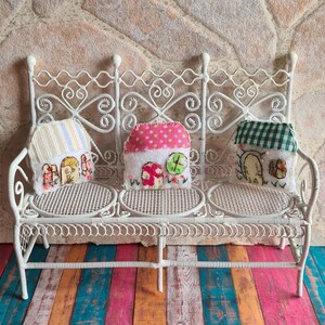 May include: Three small, handmade houses with different fabric patterns are displayed on a white, wrought iron bench. The houses are decorated with embroidered details and are sitting on a colorful wooden floor.