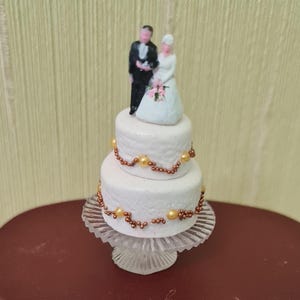 Puede incluir: Un pastel de boda blanco de dos niveles decorado con cuentas doradas y marrones. El pastel está coronado con una figura de novia y novio.
