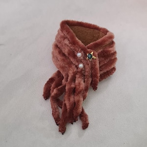 May include: A dusty rose-colored faux fur scarf with a decorative star-shaped brooch. The brooch features a green gem and gold-colored accents, along with two small, white pearl-like beads. The scarf has a soft, textured appearance.