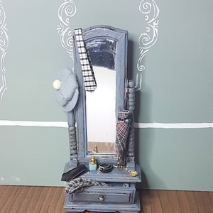 Peut inclure: Un petit miroir de coiffeuse bleu avec un tiroir, contenant une cravate à carreaux noir et blanc, un chapeau gris et une cravate à carreaux rouge. Un petit flacon de parfum et d'autres accessoires sont sur la coiffeuse. Le miroir est posé contre un mur vert clair.