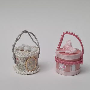 May include: Two miniature decorative hat boxes. One is white with a silver handle, floral accents, and a gold-framed oval. The other is pink with a pink bow, handle, and floral details. Both are round.