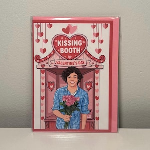 May include: Valentine's Day card with a cartoon illustration of a person holding pink roses in front of a 'Kissing Booth'. The card has a pink and red colour scheme with heart accents and the text 'Valentine's Day'.