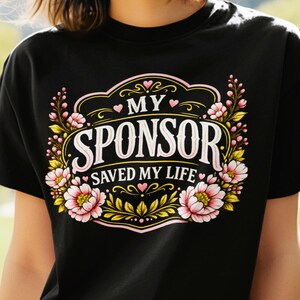 以下が含まれることがあります： 「MY SPONSOR SAVED MY LIFE」という白とピンクの文字が中央に配置された黒の半袖Tシャツ。周囲はピンクの花、緑の葉、小さなピンクのハートで構成された装飾的なフレームで囲まれています。