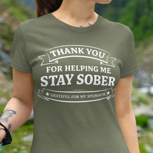 Può includere: T-shirt verde oliva con testo bianco: "THANK YOU FOR HELPING ME STAY SOBER" e "GRATEFUL FOR MY SPONSOR". La maglietta ha maniche corte e scollo tondo.
