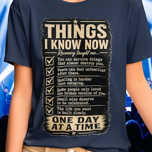 以下が含まれることがあります： 「THINGS I KNOW NOW」と「ONE DAY AT A TIME」の文字が入ったネイビーブルーのTシャツ。回復に関する声明のリストが特徴です。テキストは、使い古されたヴィンテージスタイルです。