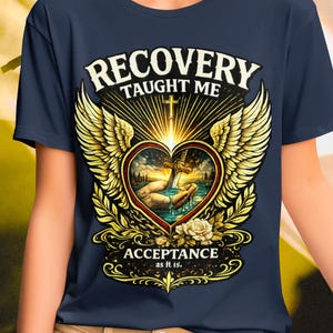 Pode incluir: Uma t-shirt azul-marinho com uma estampa gráfica de um coração com asas, uma espada e uma cruz. O texto diz RECOVERY TAUGHT ME ACCEPTANCE as it is. O design inclui raios dourados, uma mão e uma rosa branca na base do coração.