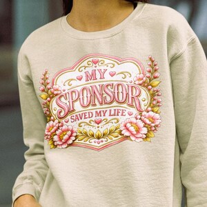 以下が含まれることがあります： ピンクの花のグラフィックプリントと装飾的なフレーム内の「MY SPONSOR SAVED MY LIFE」というテキストが特徴のベージュのクルーネックトレーナー。つばの広いピンクの帽子とダークブルーのデニムジーンズを合わせています。