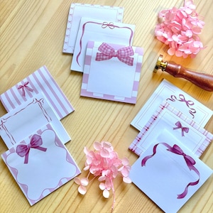 May include: Various pink and white notepads with bow designs, displayed on a wooden surface. The notepads showcase different bow styles and patterns, including stripes and gingham. A wooden handled wax stamp and pink flowers are also visible.
