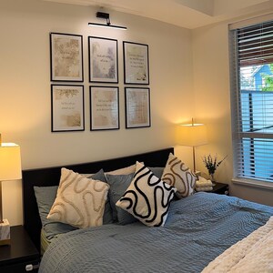 May include: A bedroom scene featuring a bed with blue bedding, decorative pillows, and a white throw blanket. Two bedside lamps with cream-colored shades flank the bed. Six framed prints hang above the bed, and a window with blinds offers a view.