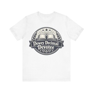 Może przedstawiać: Biały t-shirt z krótkim rękawem z ciemnoniebieską okrągłą grafiką na klatce piersiowej. Wzór zawiera otwartą książkę, trzy gwiazdki oraz wstęgę z napisem Dewey Decimal Devotee, otoczoną ozdobną obwódką z liści laurowych.
