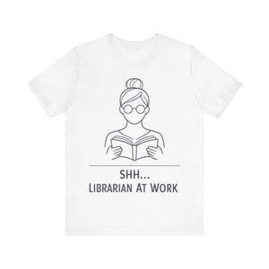 May include: A white short-sleeve t-shirt featuring a black line art graphic of a person with a hair bun and round glasses reading a book. Below the graphic, the text reads SHH... LIBRARIAN AT WORK in a simple, dark gray sans-serif font.