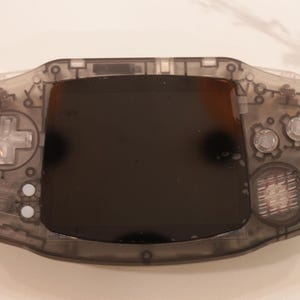 Könnte beinhalten: Eine tragbare Videospielkonsole mit einem durchscheinenden dunkelgrauen Kunststoffgehäuse. Das Gerät verfügt über einen zentralen schwarzen Bildschirm, ein Steuerkreuz auf der linken Seite, zwei runde Aktionstasten rechts und einen Lautsprecher.