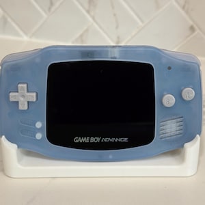 Può includere: Una console per videogiochi portatile con un guscio in plastica azzurro traslucido, uno schermo nero e pulsanti grigi. Il dispositivo è dotato di un pad direzionale, pulsanti A e B e pulsanti start e select. Il testo GAME BOY ADVANCE è stampato sotto lo schermo.