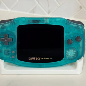 May include: A handheld Game Boy Advance video game console featuring a translucent teal plastic shell, a black screen bezel, dark gray buttons, and the text "GAME BOY ADVANCE" printed in white below the screen, resting on a white plastic stand.