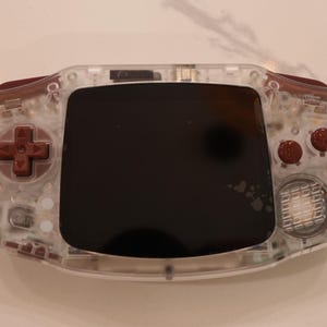 May include: A handheld video game console featuring a clear, transparent plastic outer shell. The device includes a dark screen, a maroon directional pad on the left, and two maroon action buttons on the right, with a circular speaker grille below.