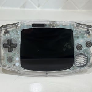 Può includere: Una console per videogiochi portatile con un guscio in plastica trasparente che rivela i componenti elettronici interni. Il dispositivo include uno schermo nero centrale, un pad direzionale grigio scuro a sinistra e due pulsanti circolari a destra.
