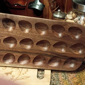 May include: A rectangular dark brown wooden serving board featuring eighteen carved egg-shaped indentations arranged in three rows of six. The board has a smooth finish with visible wood grain patterns and a handle extending from the right side.