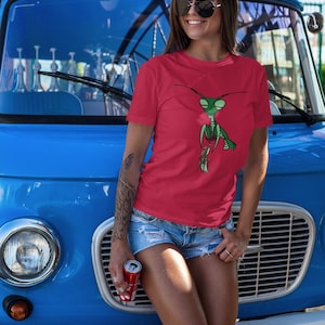 May include: Red t-shirt featuring a green cartoon praying mantis holding a red can. The person is wearing denim shorts and sunglasses, leaning against a blue vintage van. A red can is held in the person's hand.