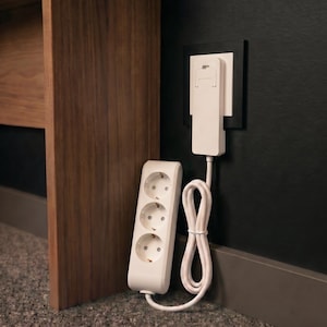 Peut inclure: Une multiprise blanche à trois prises circulaires reliée par un cordon blanc à un adaptateur mural plat et blanc. L'adaptateur est branché dans une prise murale noire, et la multiprise est posée verticalement sur le sol.