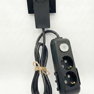 May include: A black electrical power strip with two outlets and a circular power button, connected to a black wall plug adapter. The black power cord is neatly coiled and secured with a piece of brown twine, hanging vertically from the wall plug.