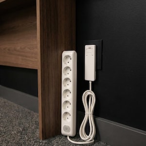 May include: A white six-outlet power strip with a circular power button at the base, connected by a white cord to a flat white plug inserted into a black wall outlet. The power strip stands vertically against a dark wall and a wooden furniture leg.