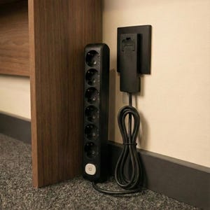 Puede incluir: Una regleta negra de seis tomas con un botón de encendido circular blanco, conectada a un adaptador de corriente negro montado en la pared con un cable negro enrollado. La regleta se apoya verticalmente junto a una pata de mueble.