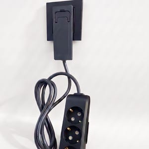 May include: A black electrical power strip with three circular outlets featuring grounding pins, connected by a black cord to a black wall plug adapter that is inserted into a black square wall plate mounted on a flat surface.