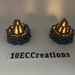 May include: Two miniature campfire decorations. Each has a ring of gray stones surrounding a pile of wooden logs with a warm, glowing light inside. The text "10ECCreations" is below the campfires.
