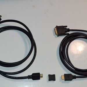 May include: Two black electronic cables are displayed. One cable features a mini-connector and a standard HDMI plug. The other cable has a DVI connector and an HDMI plug. A small, square black protective cap is positioned between the two cables.