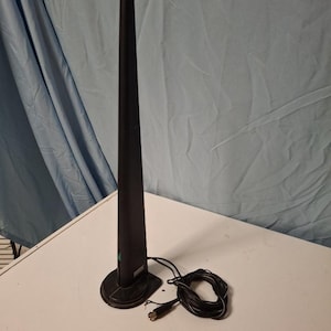 Könnte beinhalten: Eine hohe, schwarze, spitz zulaufende Fernsehantenne mit einem flachen, rechteckigen Sockel. Ein dünnes schwarzes Koaxialkabel ist neben dem Sockel aufgewickelt und endet in einem standardmäßigen Metallanschluss. Die Antenne hat eine matte Oberfläche und einen kleinen türkisfarbenen Akzent.