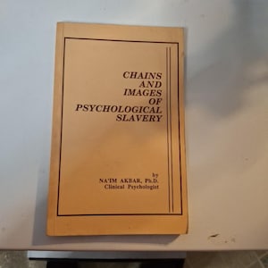 May include: A book titled "Chains and Images of Psychological Slavery" by Na'im Akbar, Ph.D., Clinical Psychologist. The book has a tan cover with black text and a black border. The book is on a white surface.