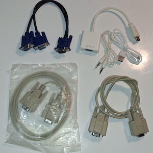May include: A collection of computer cables and adapters including a black VGA splitter cable, a white multi-port adapter with attached audio and USB cables, and two beige serial cables, one of which is sealed in a clear plastic bag.