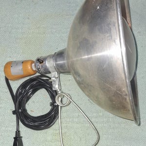 May include: Vintage metal spotlight with a large, cone-shaped reflector. It features a wooden handle, a black power cord, and a metal stand. The spotlight is made of brushed metal and has a classic design.