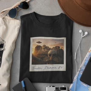 May include: Black t-shirt featuring a retro-style image of aliens visiting Mount Rushmore, with the text "South Dakota 89'". The image includes a UFO and a vintage polaroid frame. The shirt is surrounded by accessories.