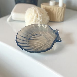 以下が含まれることがあります： 半透明の青い貝殻型の石鹸皿。皿にはスカラップエッジとテクスチャード加工された内側があります。他のバスルームアクセサリーが背景にあります。