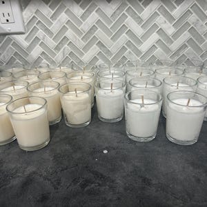 Puede incluir: Una colección de pequeñas velas votivas de vidrio transparente llenas de cera blanca. Cada vela tiene una sola mecha centrada en la cera. Las velas están dispuestas sobre una encimera oscura frente a un salpicadero claro con dibujos.