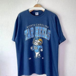 May include: A navy blue t-shirt featuring the words "North Carolina Tar Heels" in light blue arched lettering. Below the text is a cartoon ram mascot in a light blue uniform. The shirt is hanging on a white wall.