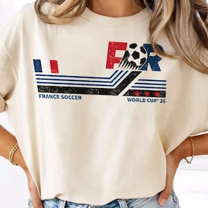Puede incluir: Una camiseta de color crema con un gr&aacute;fico desgastado que incluye el texto FRANCE SOCCER y WORLD CUP '26. El dise&ntilde;o presenta un bal&oacute;n de f&uacute;tbol, rayas azules y rojas, y tres estrellas rojas, todo en un estilo vintage y envejecido.