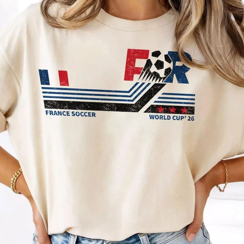 Puede incluir: Una camiseta de color crema con un gr&aacute;fico desgastado que incluye el texto FRANCE SOCCER y WORLD CUP '26. El dise&ntilde;o presenta un bal&oacute;n de f&uacute;tbol, rayas azules y rojas, y tres estrellas rojas, todo en un estilo vintage y envejecido.
