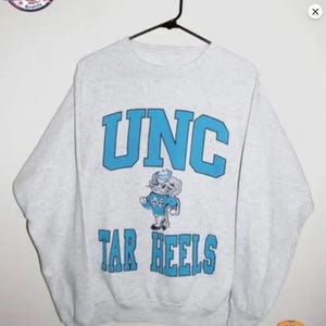 May include: A light gray sweatshirt with the text "UNC" in light blue block letters. Below the text is a cartoon mascot in a light blue outfit. The text "TAR HEELS" is below the mascot in light blue block letters.