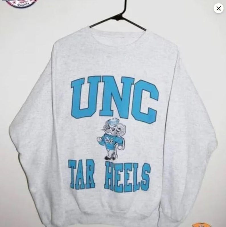 May include: A light gray sweatshirt with the text "UNC" in light blue block letters. Below the text is a cartoon mascot in a light blue outfit. The text "TAR HEELS" is below the mascot in light blue block letters.