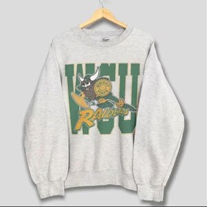 May include: A light grey sweatshirt with a crew neck and long sleeves. The front features a green and yellow graphic of a buffalo with the text "WSU RAIDERS". The sweatshirt is hanging on a wooden hanger.