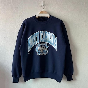 Puede incluir: Sudadera azul marino con la inscripci&oacute;n "NORTH CAROLINA" en azul claro arqueada sobre el pecho. Debajo, un logotipo azul claro con "CAROLINA TAR HEELS". La sudadera est&aacute; colgada de una percha de madera.